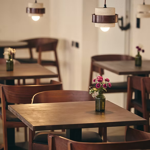 Empty cafe interior with wooden tables and chairs, warm pendant lights overhead, and a small vase of pink flowers on the closest table.