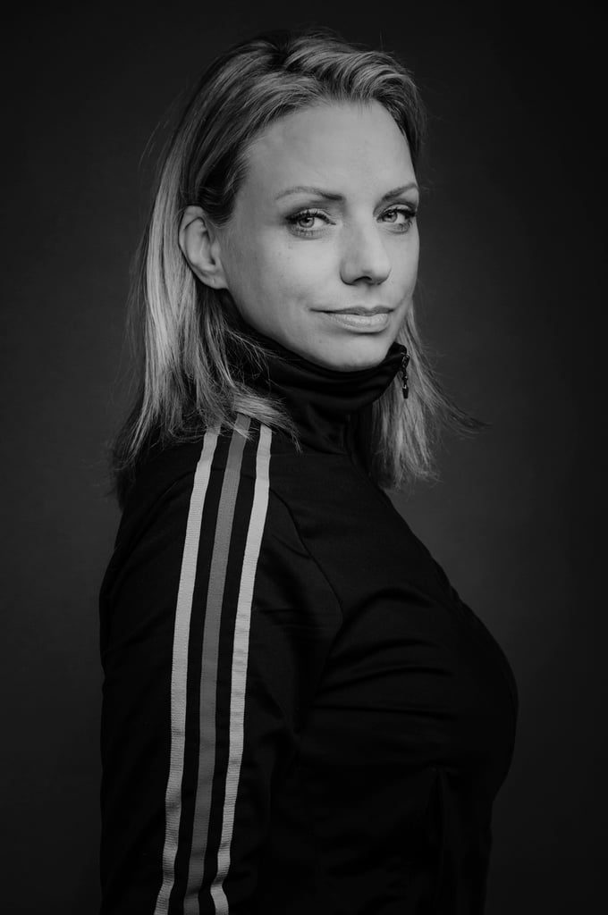 Black-and-white portrait of a woman wearing a sporty jacket with three stripes, looking slightly over her shoulder at the camera.