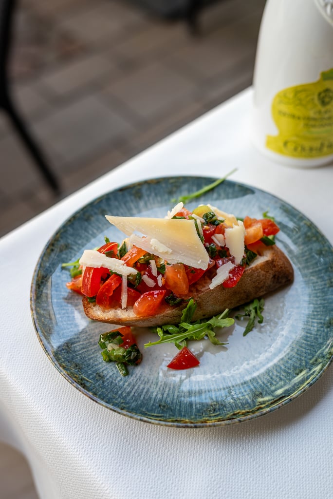 Slice of bruschetta on a blue plate topped with diced tomatoes, herbs, and cheese shavings with greens nearby on a white tablecloth.