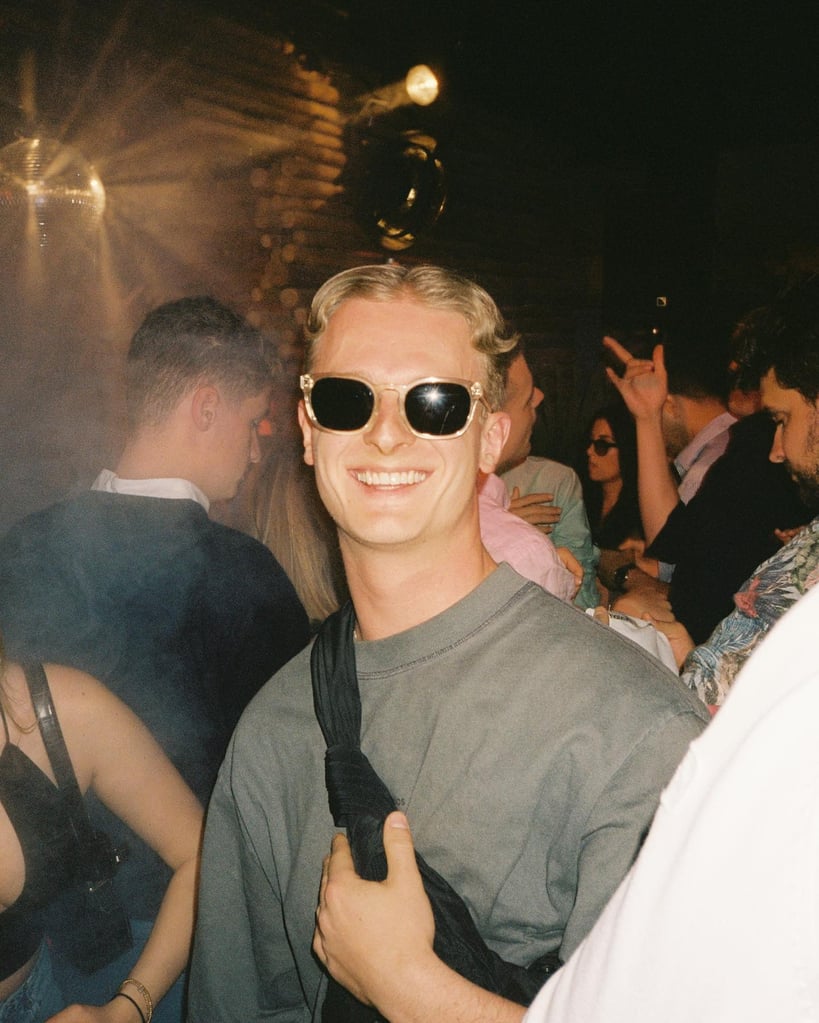 Smiling man with blond hair and sunglasses at a crowded nighttime party, wearing a gray shirt and crossbody strap, lit by warm lights.