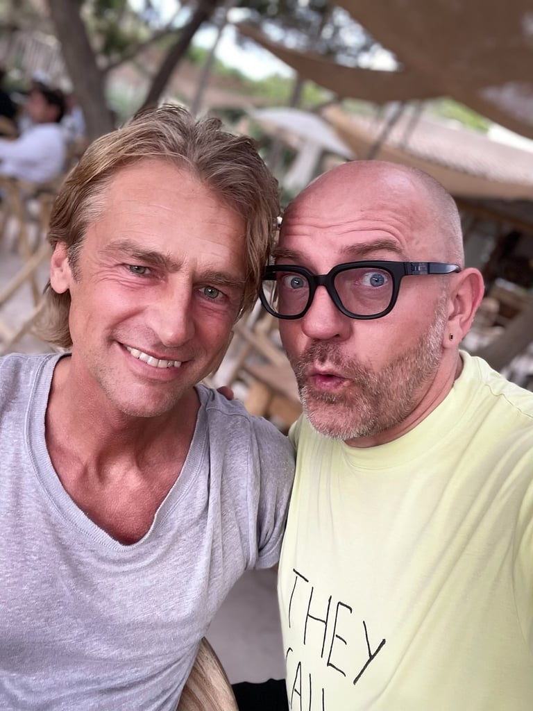 Two men pose for a selfie outdoors, smiling at the camera with a blurred cafe-style background behind them.