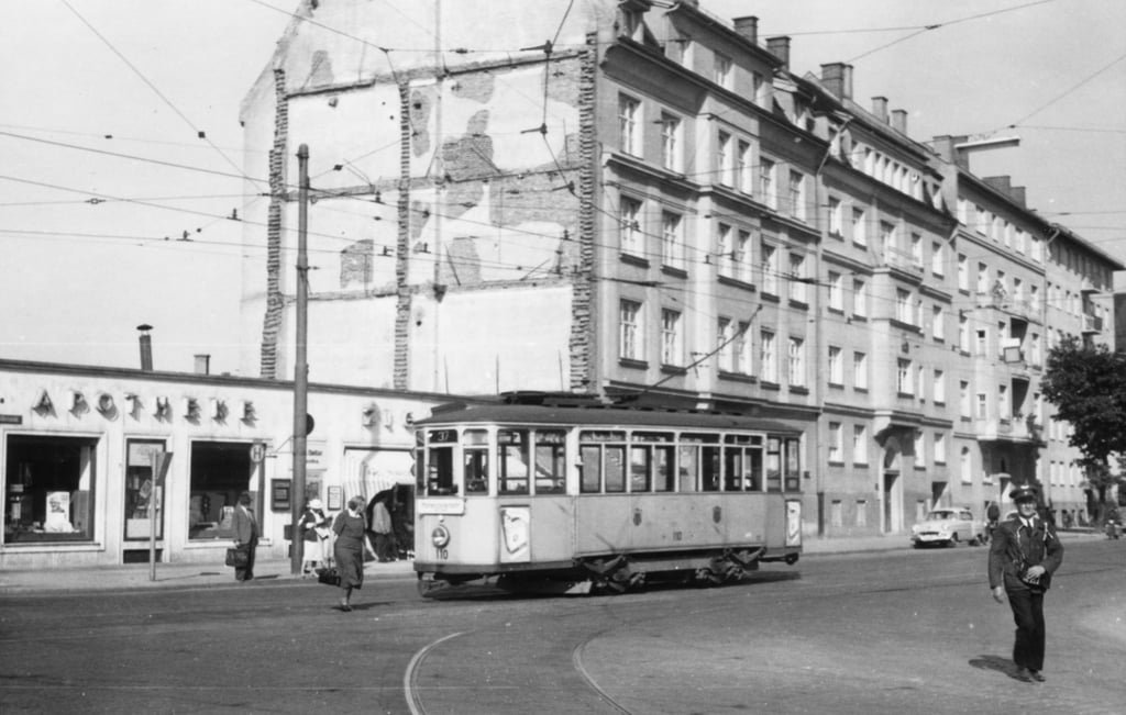 A streetcar runs along tracks in a city street, with pedestrians nearby and a row of multi-story buildings in the background.