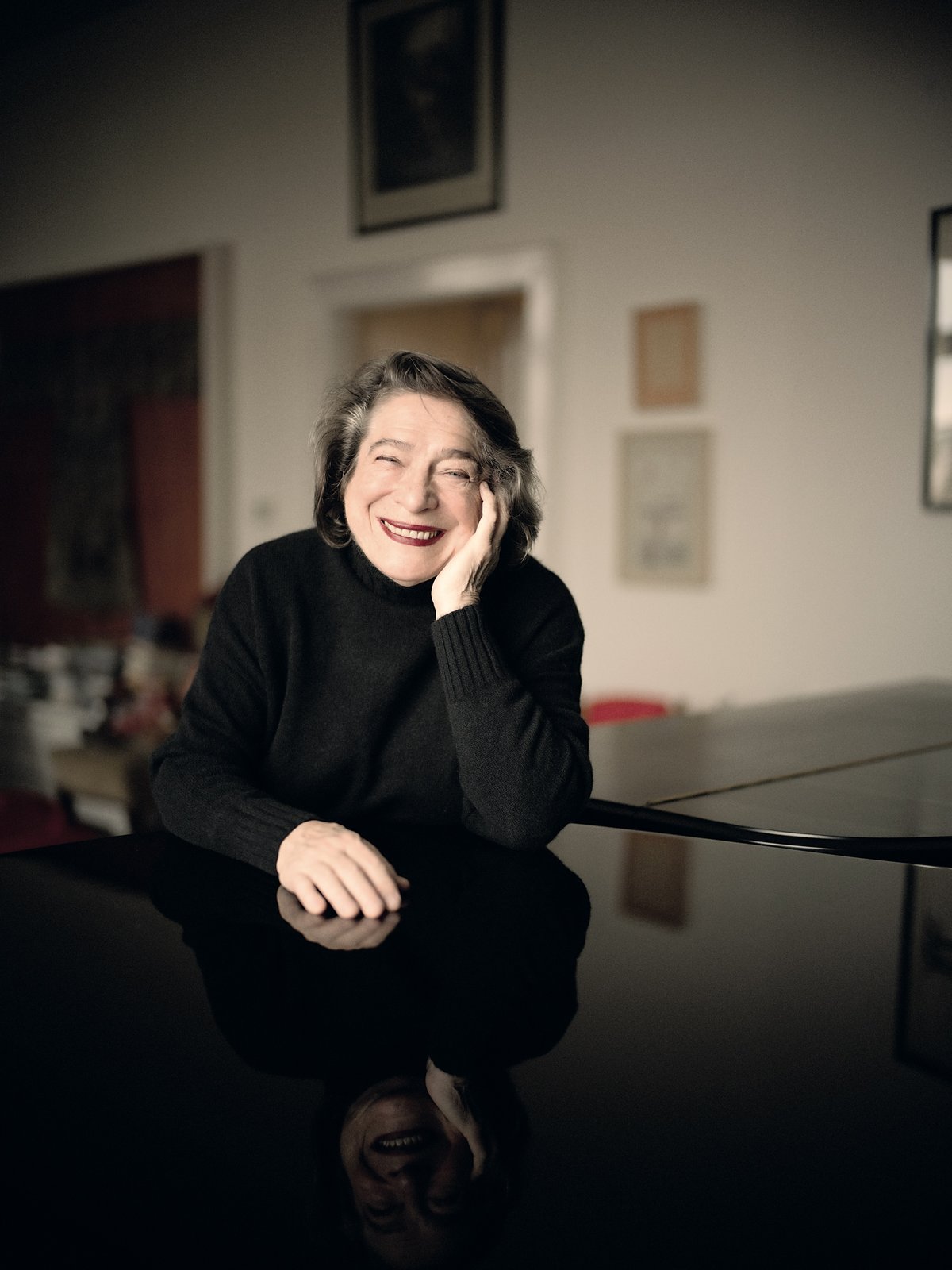 Smiling elderly woman in a black sweater leaning on a piano with her head resting on her hand, indoors, reflection on the piano surface shows her face.