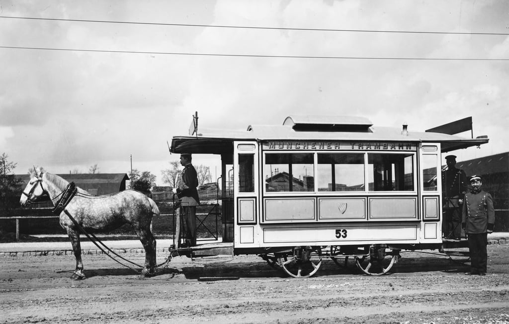 Black-and-white photo of a horse-drawn streetcar, number 53, labeled Münchener Trambahn, with two uniformed attendants nearby on a dirt road.