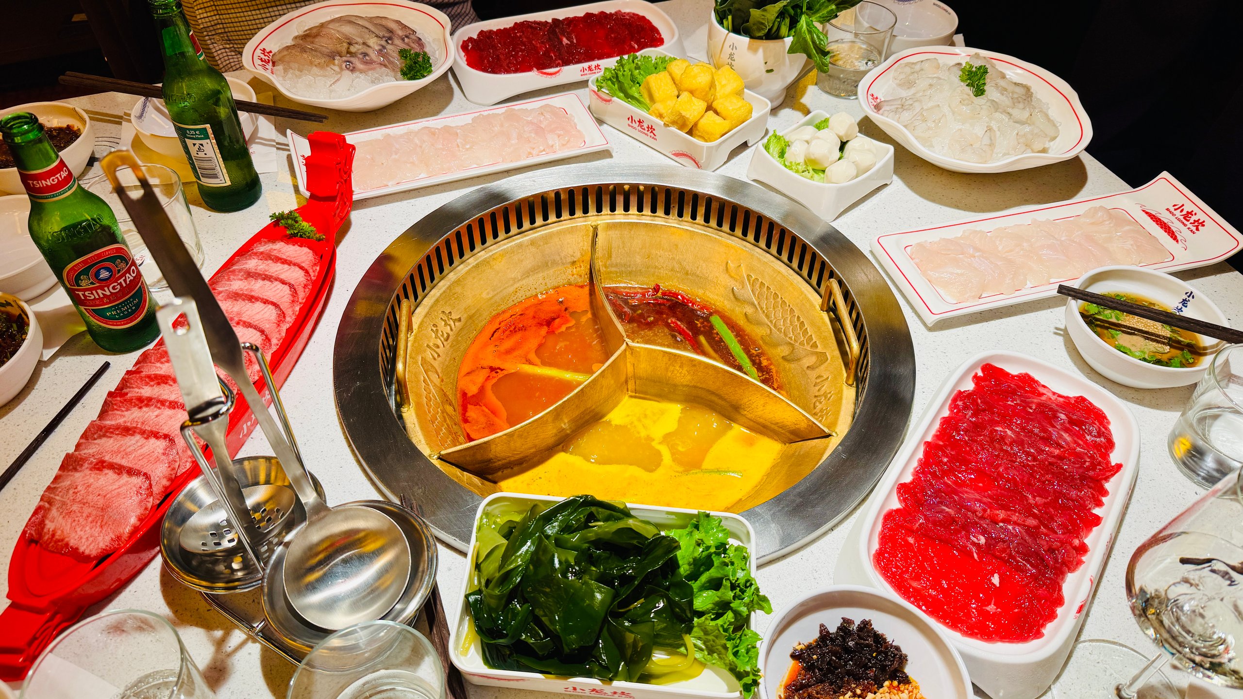 Hot-Pot-Tisch mit dreigeteiltem Topf aus roter, gelber und klarer Brühe, umgeben von Fleischplatten, Gemüse und Tsingtao-Bier