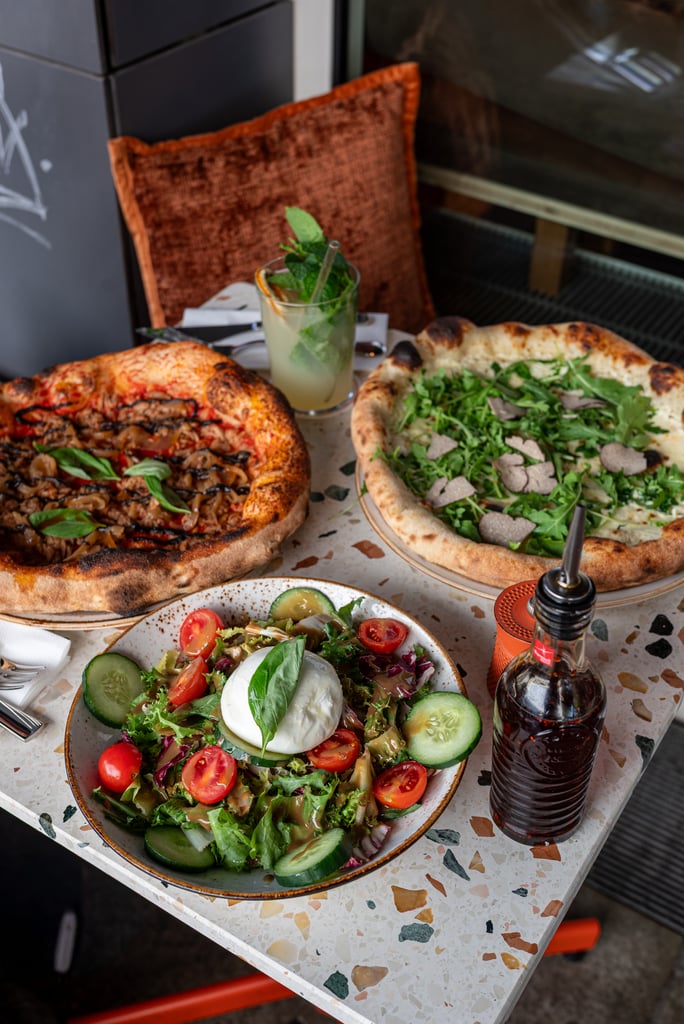 Two pizzas on a speckled table with a bowl of salad, a mint cocktail, and a bottle of soda nearby.