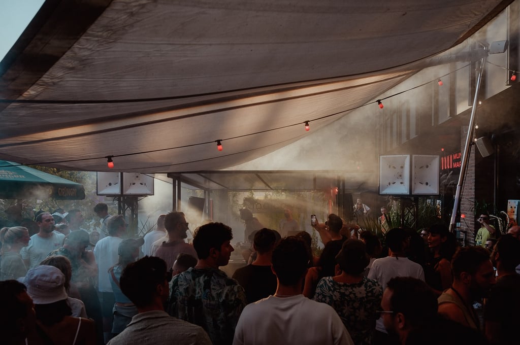 Crowd of people at an outdoor party under a large tent, with stage smoke and warm string lights in the background.