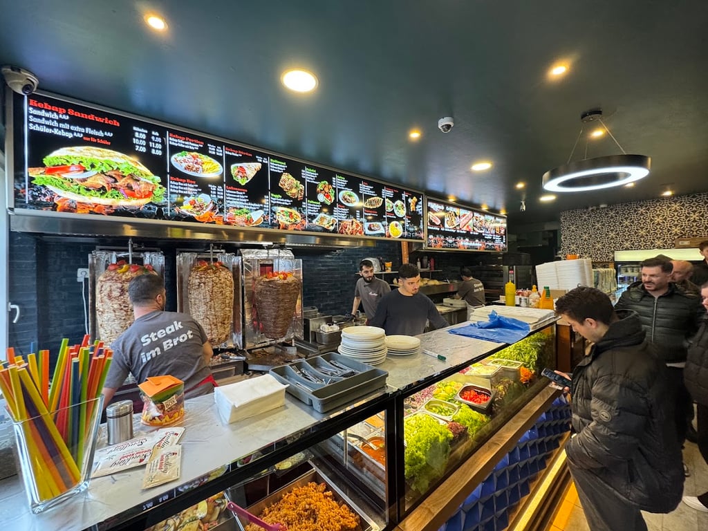 Kebab shop interior with vertical rotisserie grills, bright menu boards overhead, and staff preparing food behind the counter
