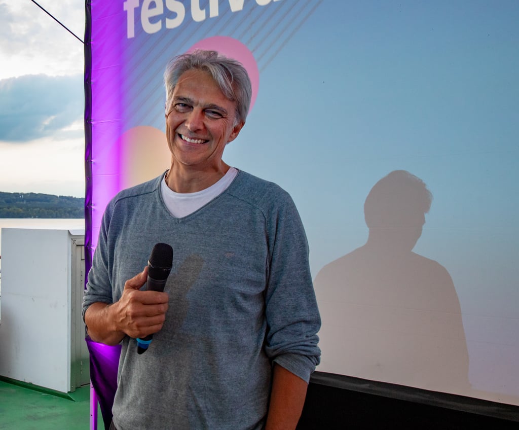 Smiling man in a gray sweater holding a microphone outdoors, with a purple banner in the background.