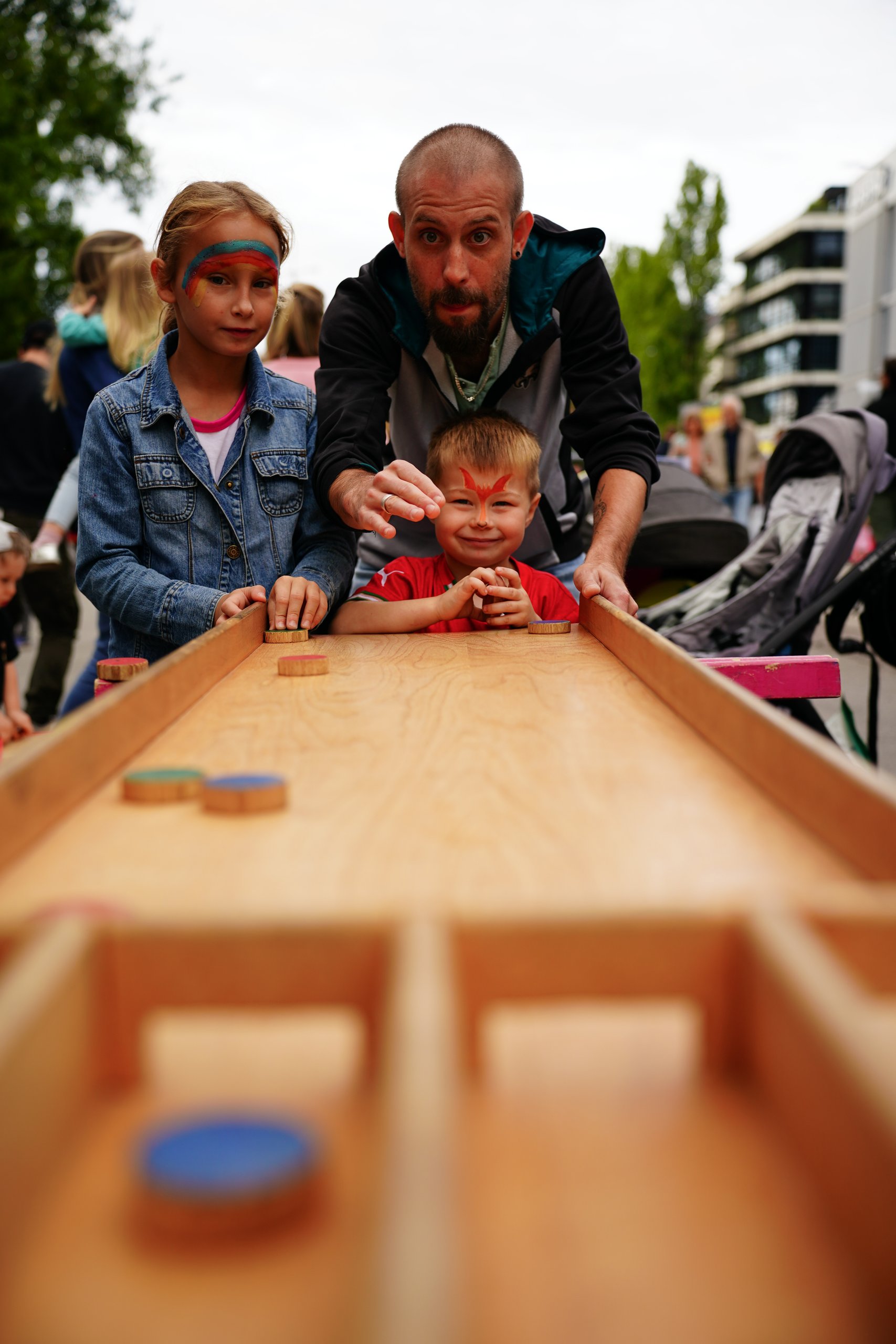 Father and two children lean over a wooden table, placing round discs in a field-game at an outdoor event; girl wears a denim jacket and rainbow face paint, boy has red face paint.