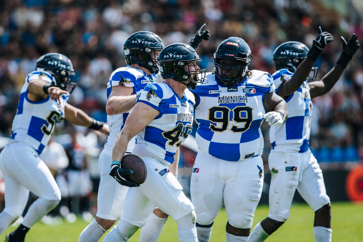 Ravens Munich rugby players in blue-and-white uniforms celebrate on the field with a cheering crowd in the background.