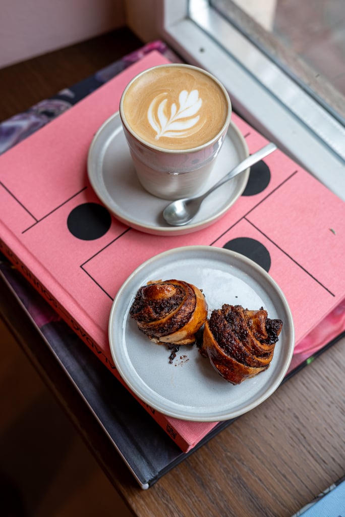 Cup of coffee with leaf-shaped latte art in a to-go cup on a metal saucer, beside a plate of cinnamon rolls on a pink box near a window.