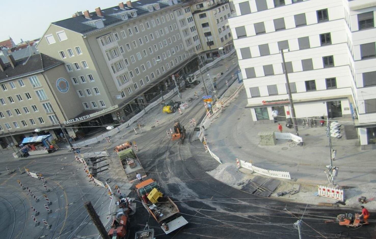 Bird's-eye view of a city street under construction with tram tracks, barriers, and construction equipment between beige and white buildings outside a cafe.