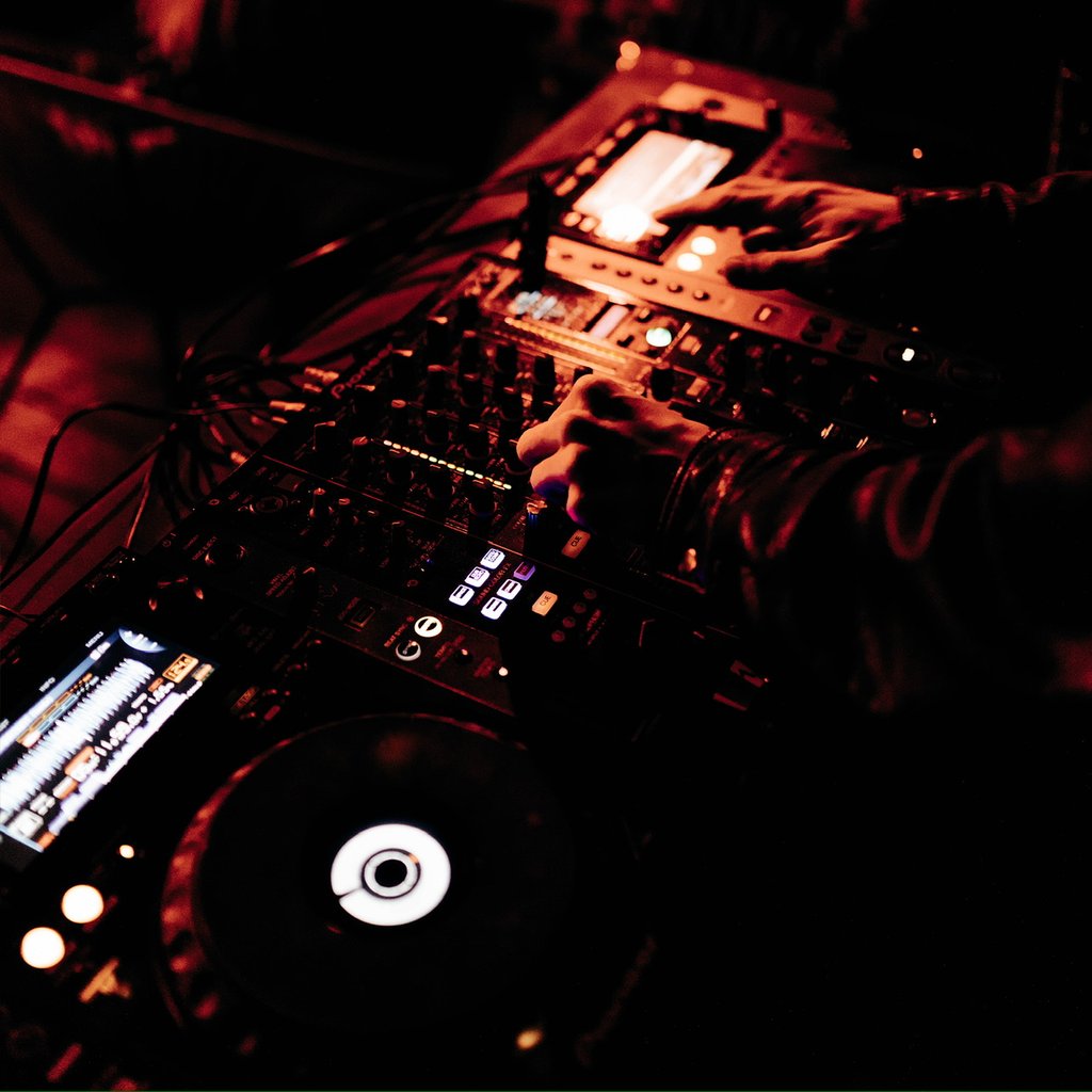 DJ performing on a dark red-lit setup, hands turning knobs on a mixer with turntable and screens glowing nearby