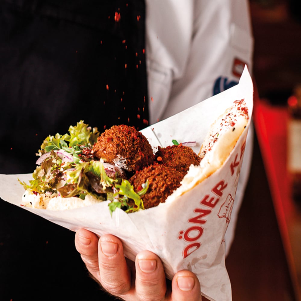 Hand holding a paper cone filled with crispy falafel balls, greens, and pita at a street-food stall.