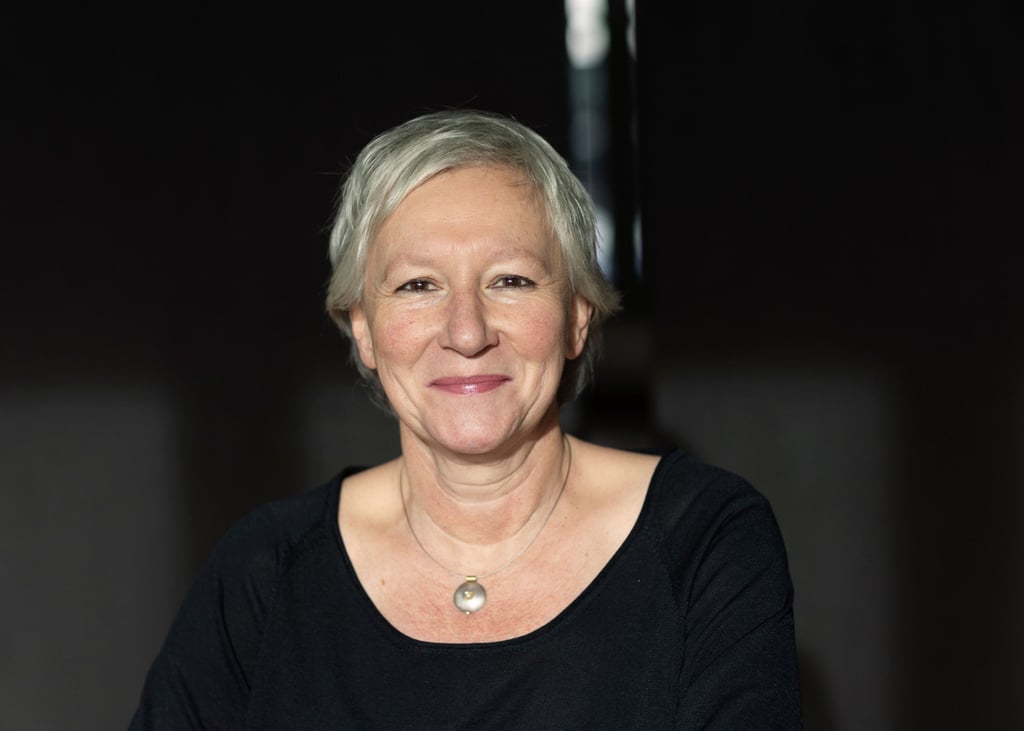Portrait of a smiling woman with short gray hair wearing a black top and a round pendant necklace against a dark background.