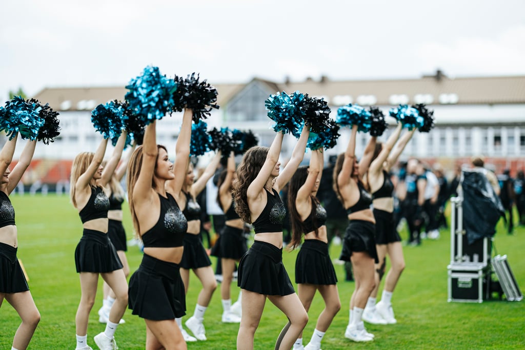 Cheerleaders in black outfits perform with blue and black pom-poms on a grassy field at an outdoor event.
