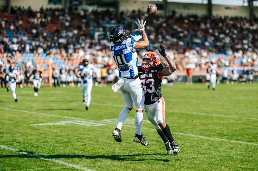 Blue-and-white jersey player Okonkwo leaps to catch a football as a defender reaches up, with a packed stadium in the background.