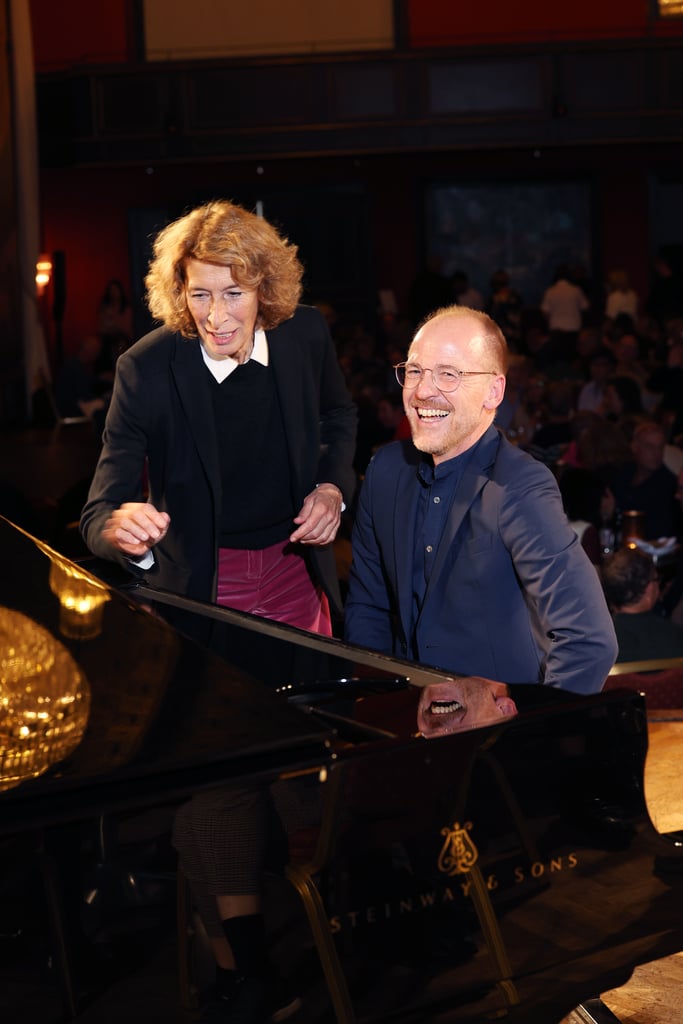 Two musicians at a grand piano during a performance, woman standing and smiling beside the pianist as the audience listens in a dimly lit venue.