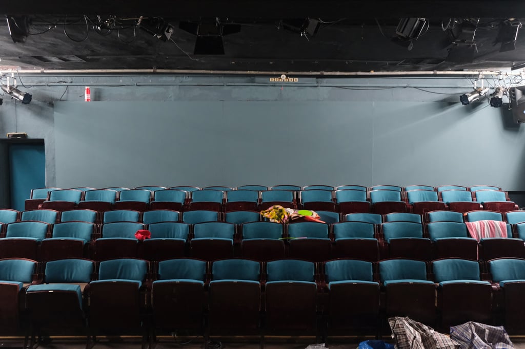 Empty theater auditorium with teal seats facing a bare stage; a few jackets and fabrics draped on seats in the middle rows.