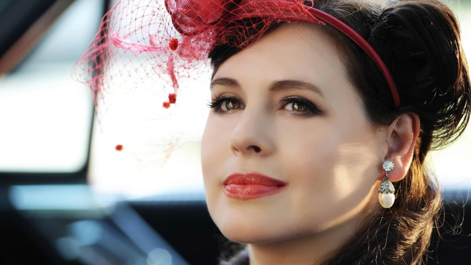 Close-up of a woman wearing a red fascinator with netting and pearl earrings, gazing softly ahead.