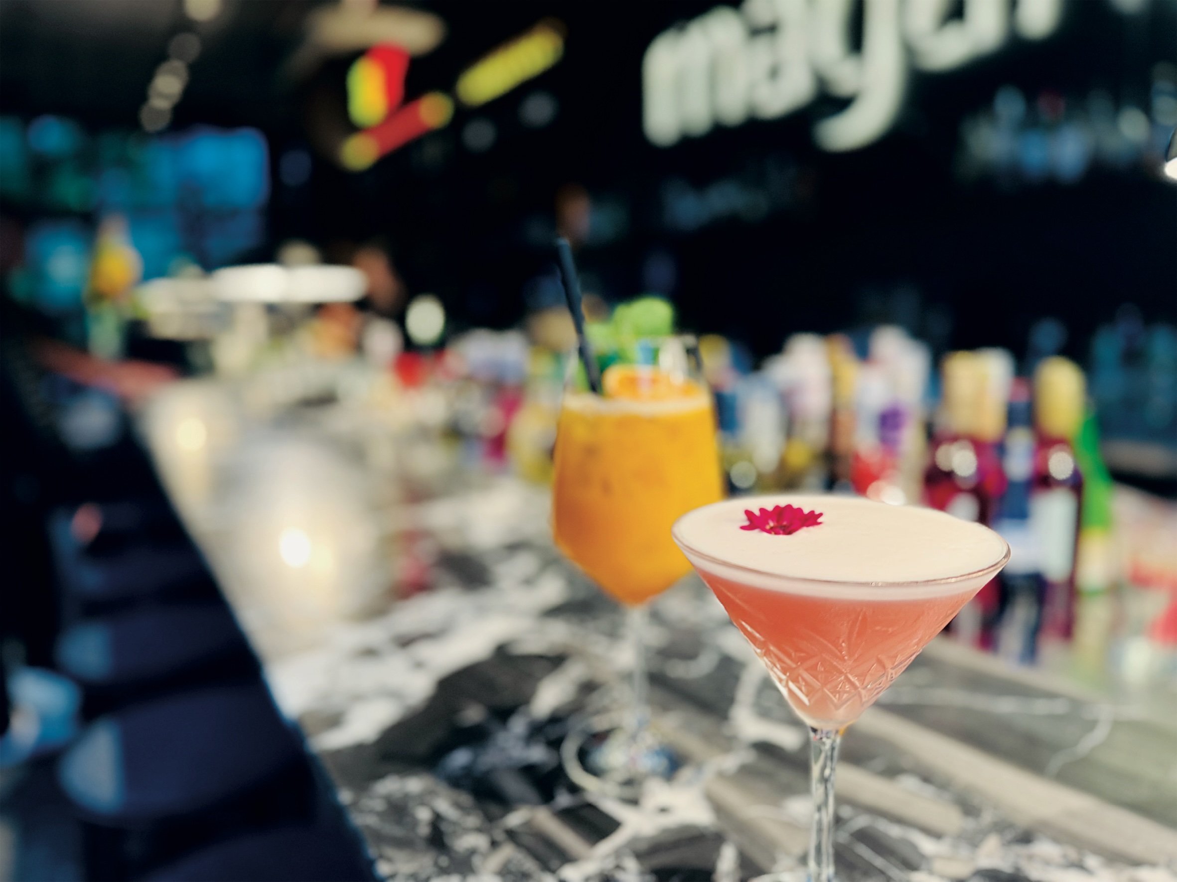 Zwei Cocktails, ein orangefarbener und ein rosa mit Blüte, auf der Marmorbar vor dem Magari-Schriftzug