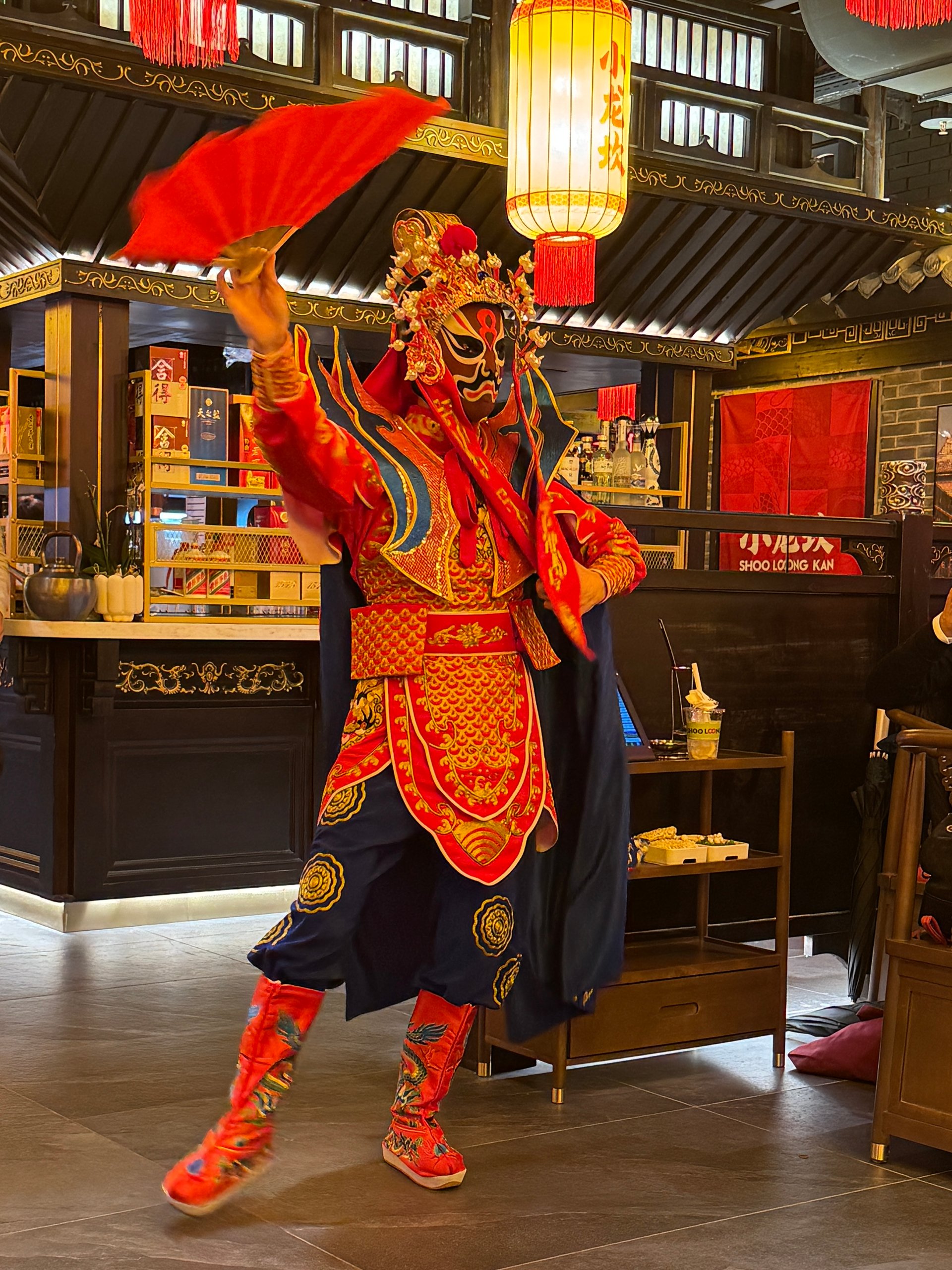 Peking opera performer in ornate red and gold costume with a painted mask waving a red fan indoors under lanterns.