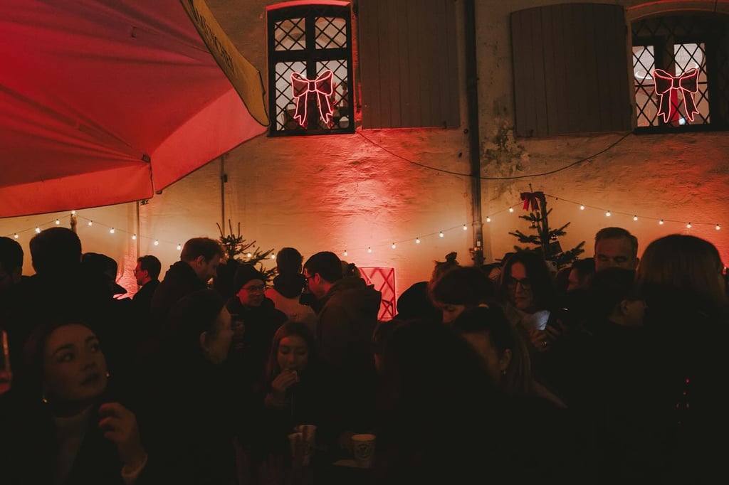 Crowd of people mingling at a holiday party in a warmly lit courtyard, with red lighting and string lights overhead.