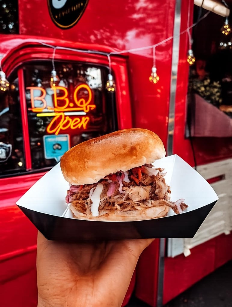 Pulled pork sandwich with onions in a paper tray held up in front of a red BBQ food truck with neon 'Open' sign.