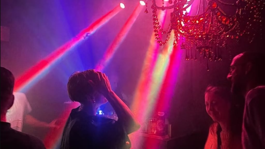 People dancing in a dim club bathed in red, blue and purple beam lights with a chandelier overhead, creating a party atmosphere.