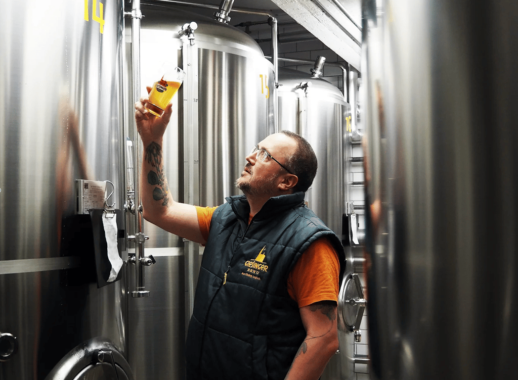 Brewing worker in a brewery holding a glass of beer up against large stainless steel fermentation tanks behind him.