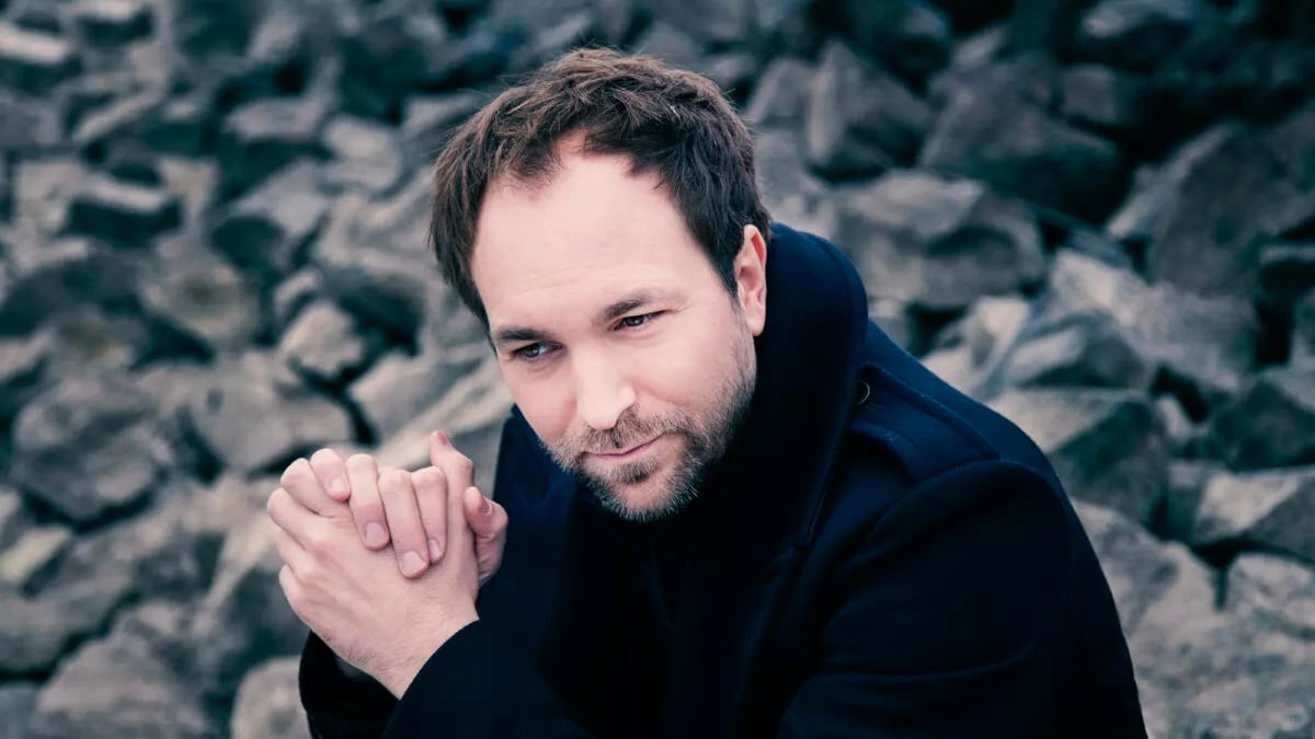 Man with short brown hair and stubble sits among gray rocks, hands clasped, looking contemplative. ETA: image shows a thoughtful pose outdoors with a rocky background.