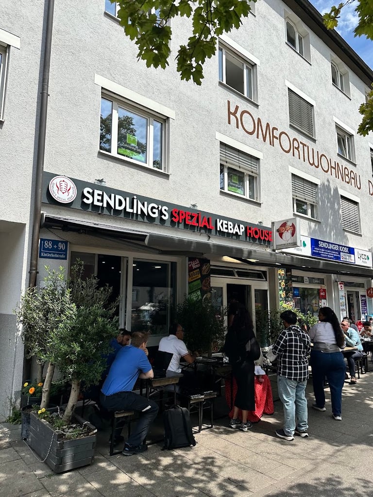 Outdoor seating in front of a kebab shop called 'Sendling's Spezial Kebap House' on a sunny street, customers sit at tables under a canopy with potted plants nearby.
