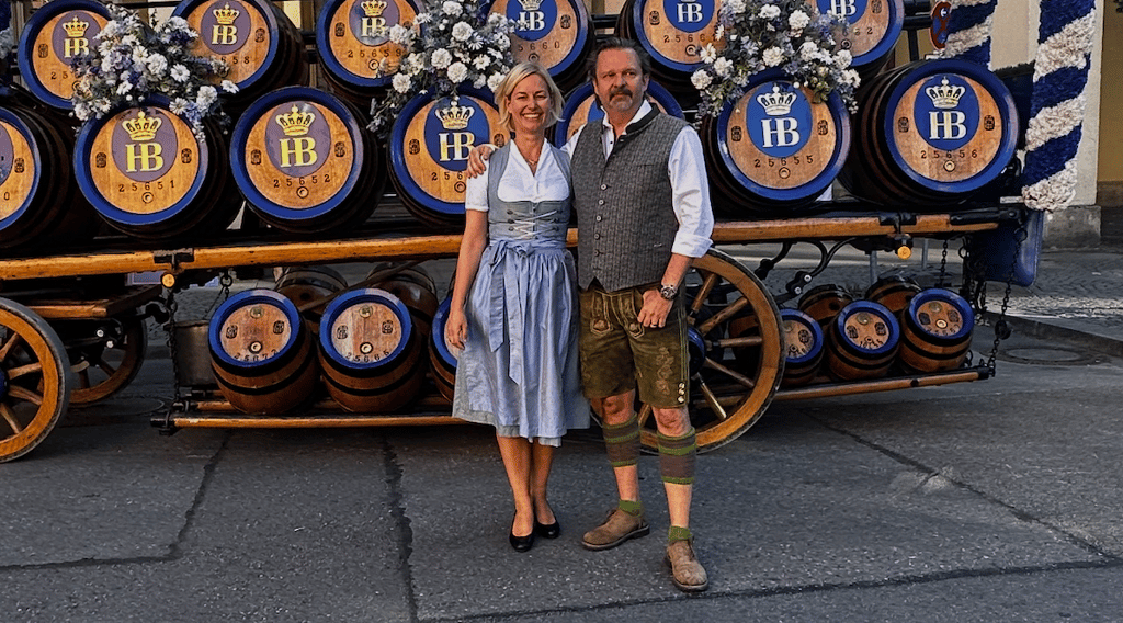 Couple in traditional Bavarian clothing posing in front of a beer wagon with blue-and-gold HB barrels and floral decorations.