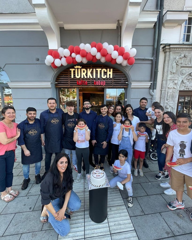 Group of people posing outside a kebab shop named Turkich under a red-and-white balloon arch, with a cake displayed in front.
