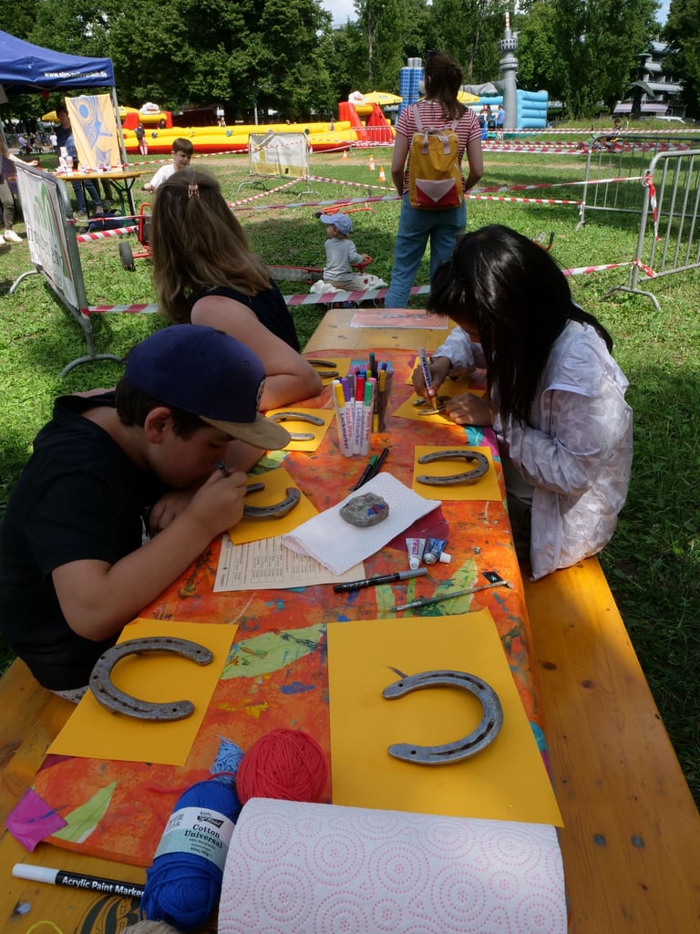 Children sitting at a colorful outdoor craft table, drawing and painting horseshoe shapes on yellow sheets, with supplies nearby and inflatable slides in the background.