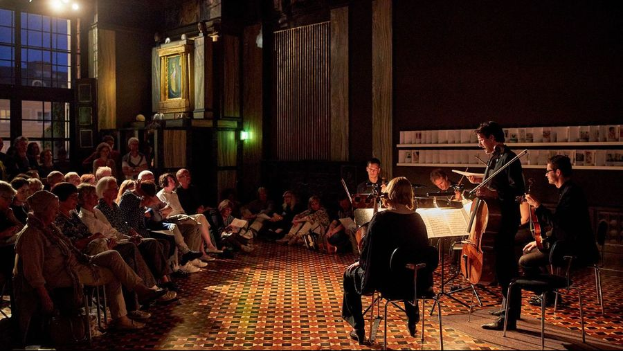 A small chamber ensemble performs on a stage in a dimly lit room while an audience sits along both sides of a patterned tile floor, listening intently.
