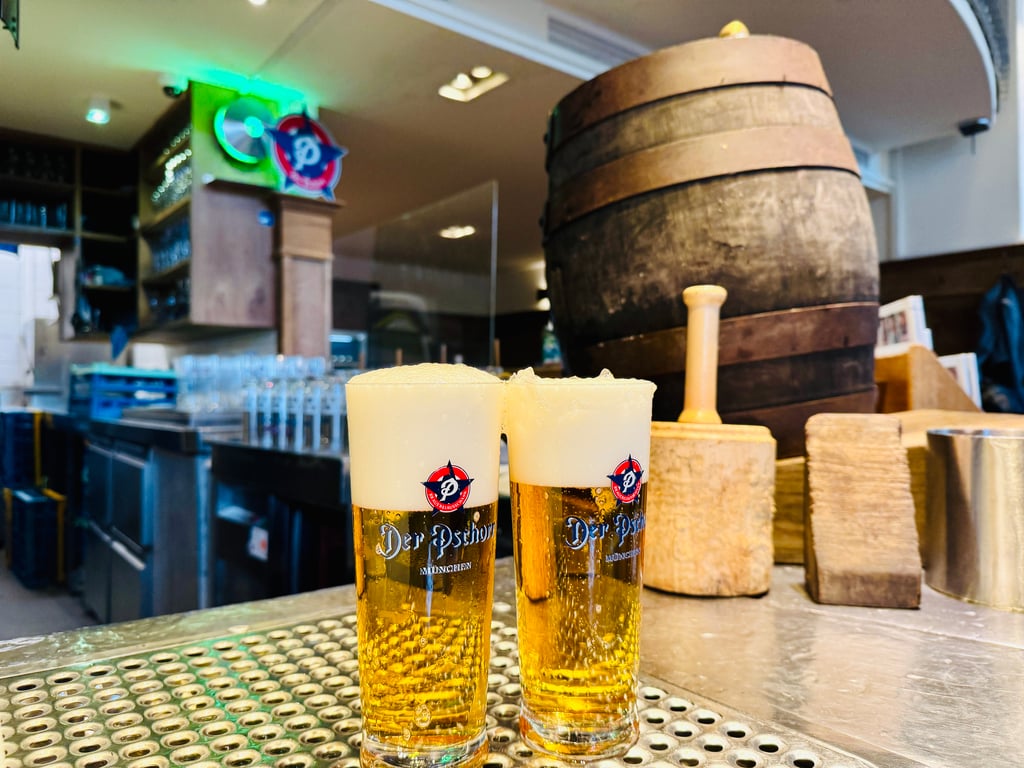 Two beer glasses with foamy heads on a metal bar, Der Pschorr logo visible, with a large wooden barrel in the background of a brewery.