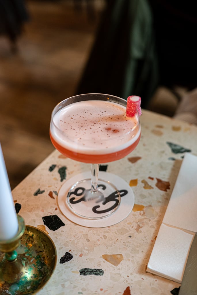 Pink frothy cocktail in a coupe glass with a pink sugared rim resting on a terrazzo table, with a round coaster underneath and a white napkin beside it.
