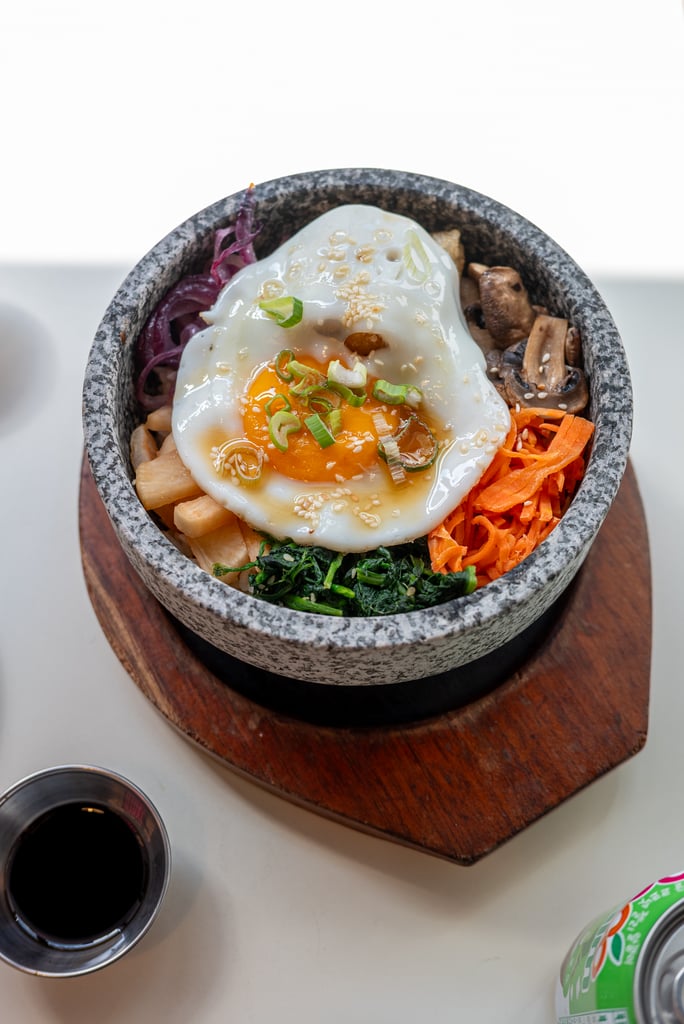 Korean bibimbap in a stone bowl topped with a fried egg, sesame seeds, and colorful vegetables like carrots, mushrooms, and greens on a wooden board.