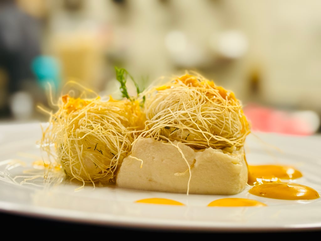 Close-up of a pastry topped with crispy shredded strands and orange sauce dots on a white plate, blurred background outdoors/restaurant setting.