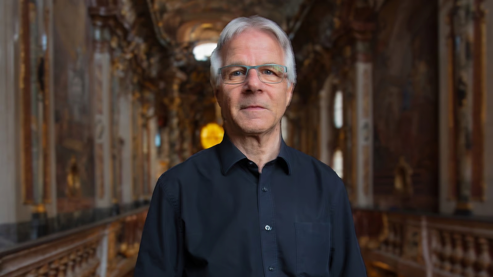 Portrait of an older man with gray hair and glasses, wearing a dark button-down shirt, standing in a richly decorated interior.