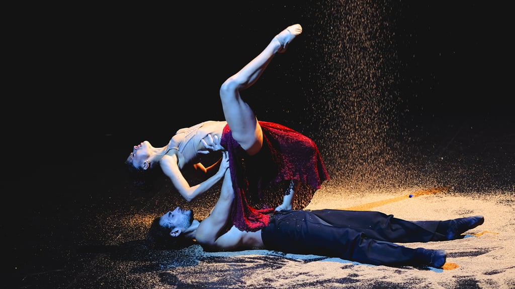 Two dancers on a dark stage: man lying on his back, woman arching above him, sand dust rising around them.