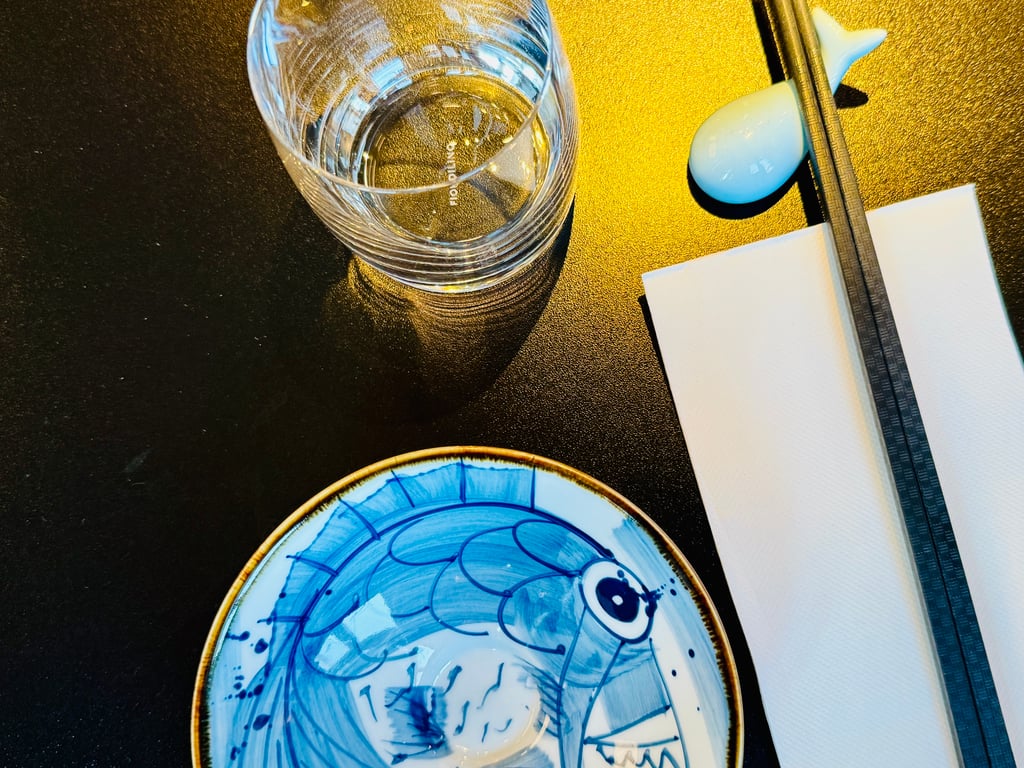 Close-up of a blue ceramic plate with wave patterns, a glass of water, a ruler, and white napkins on a dark tabletop.