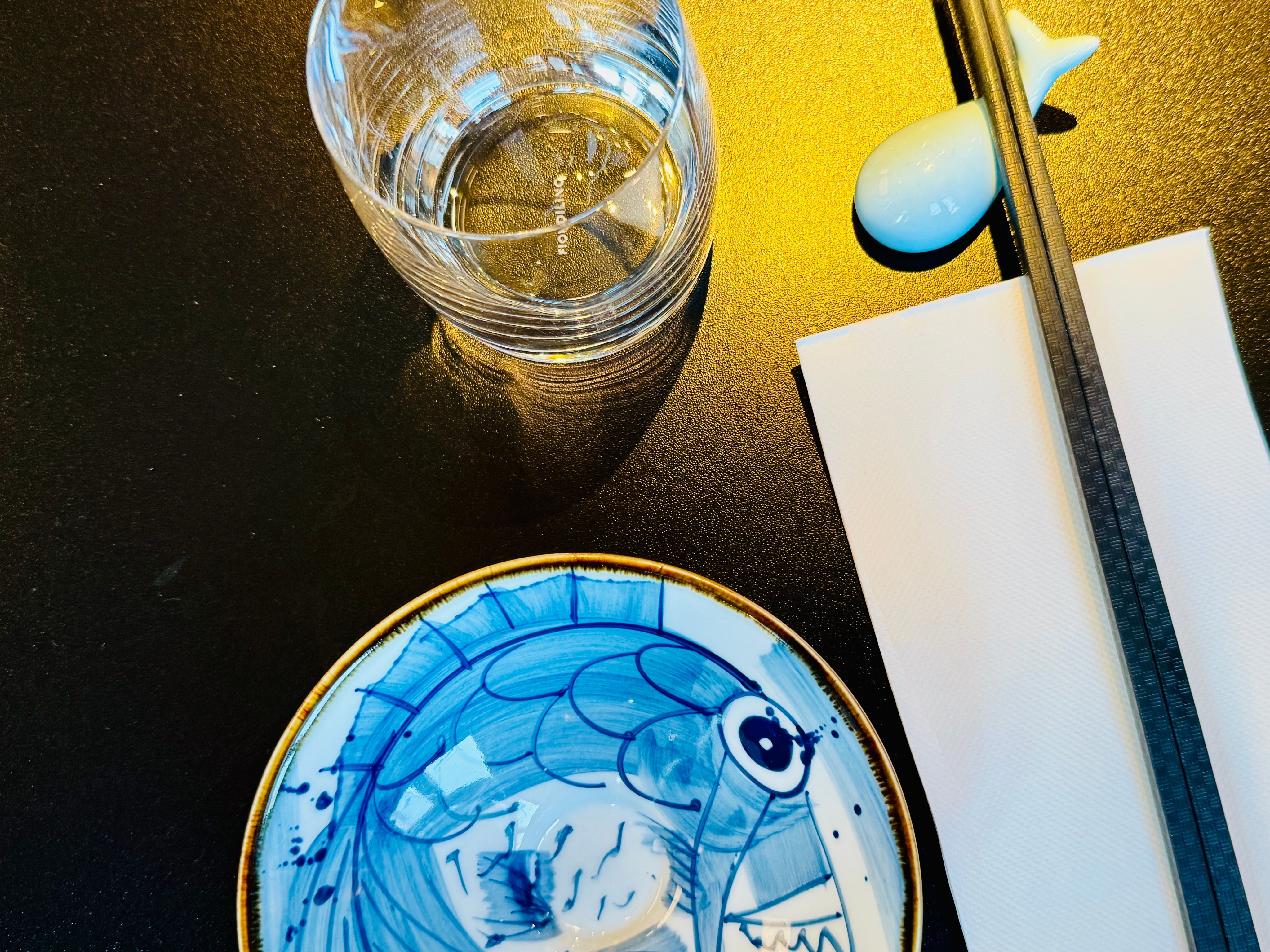 Blauer Teller mit Fischmotiv, Stäbchenablage in Fischform und Wasserglas auf einem dunklen Tisch