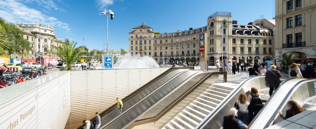 Crowded urban plaza with a sloping escalator, a central fountain spraying water, and historic beige buildings lining the square under a blue sky.