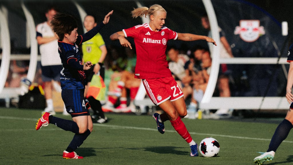 Female soccer player in a red kit with Allianz logo dribbles the ball as a blue-jersey opponent closes in on a green field.