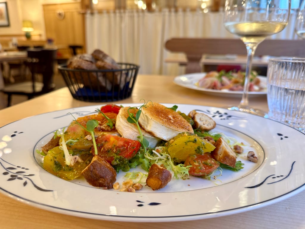 Plate of salad with tomatoes, greens, croutons and herbs on a decorative plate; blurred wine glass and bread basket in the background.