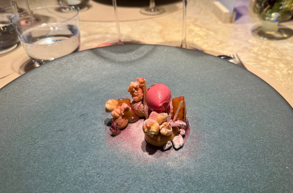Small plated dessert with pink sorbet, caramel-topped decorations, and powdered sugar on a dark round plate at a fine-dining table setting.