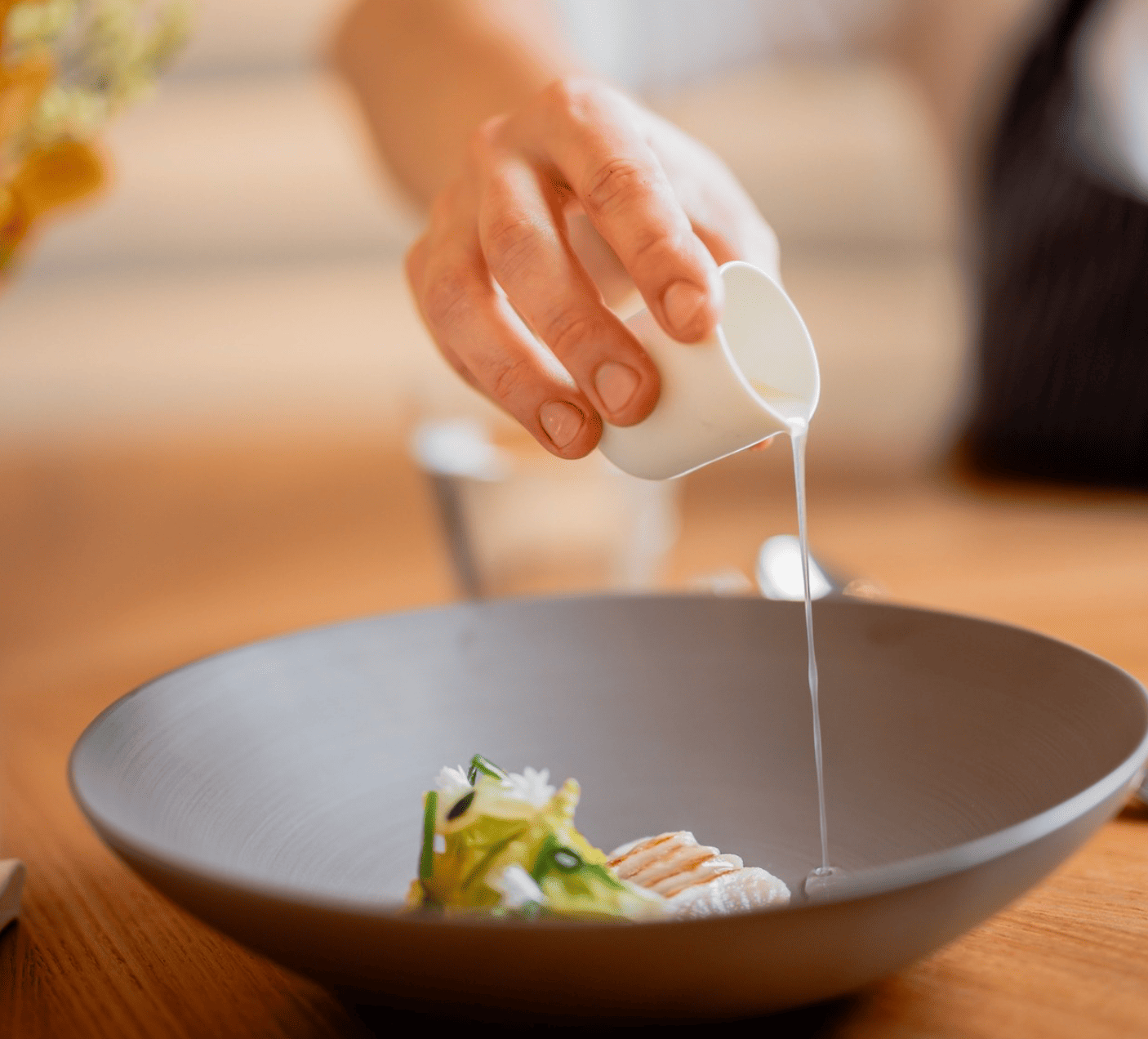 Hand gießt Sauce über ein Gericht in einer grauen Schale